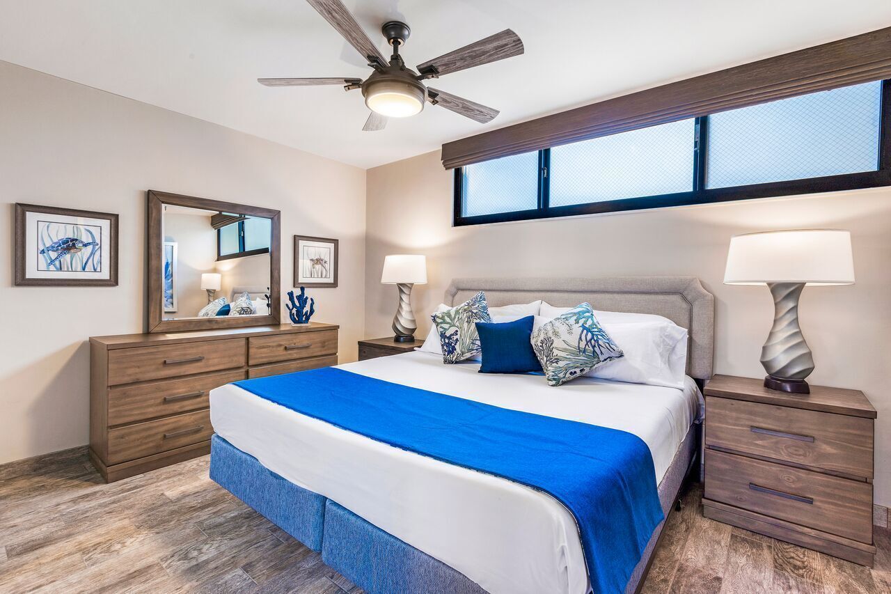 A bedroom with a large bed, a ceiling fan, a dresser, bedside lamps, and artwork on the walls.