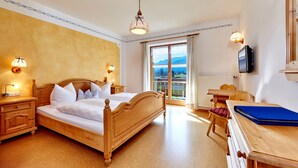A bedroom with a wooden bed, bedside lamps, a television, and a view of mountains through a large window. - Bauernhof Buchwieser (Unterammergau)