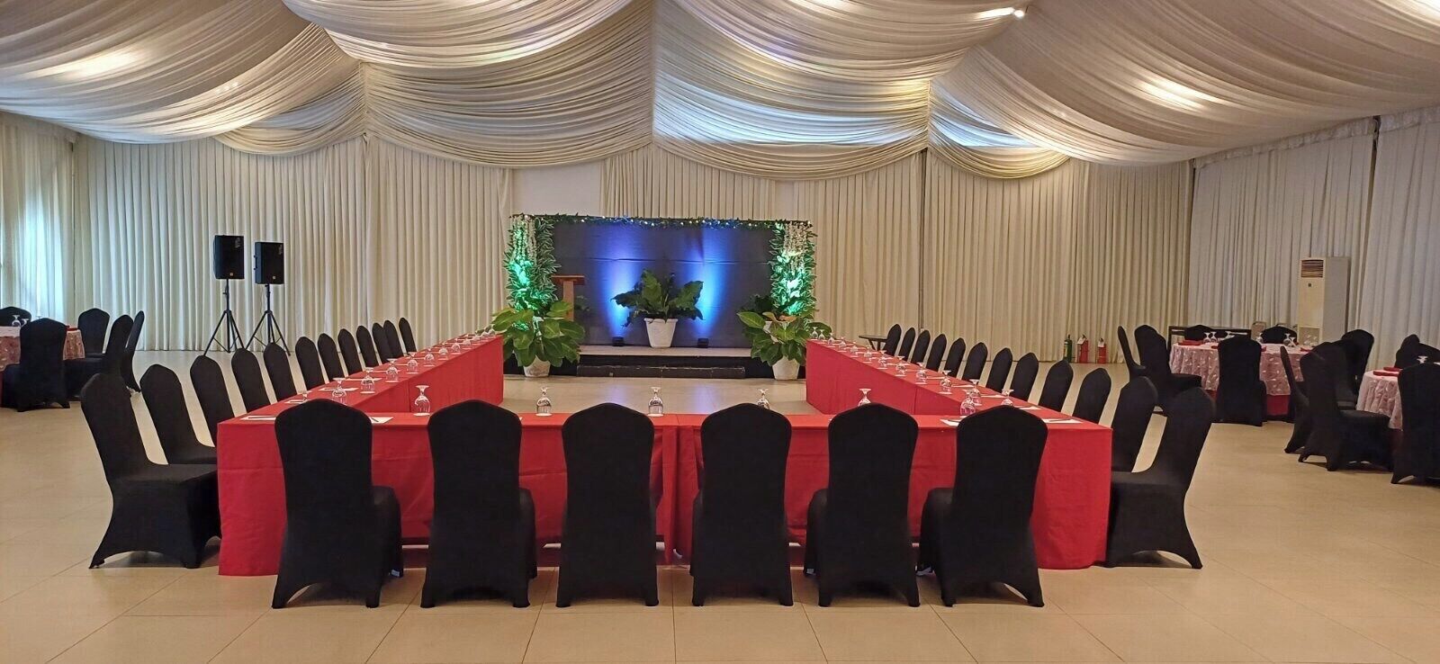 Un salón de conferencias con una larga mesa, sillas negras y un escenario con plantas e iluminación.