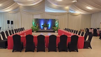 Un salón de conferencias con una larga mesa, sillas negras y un escenario con plantas e iluminación.