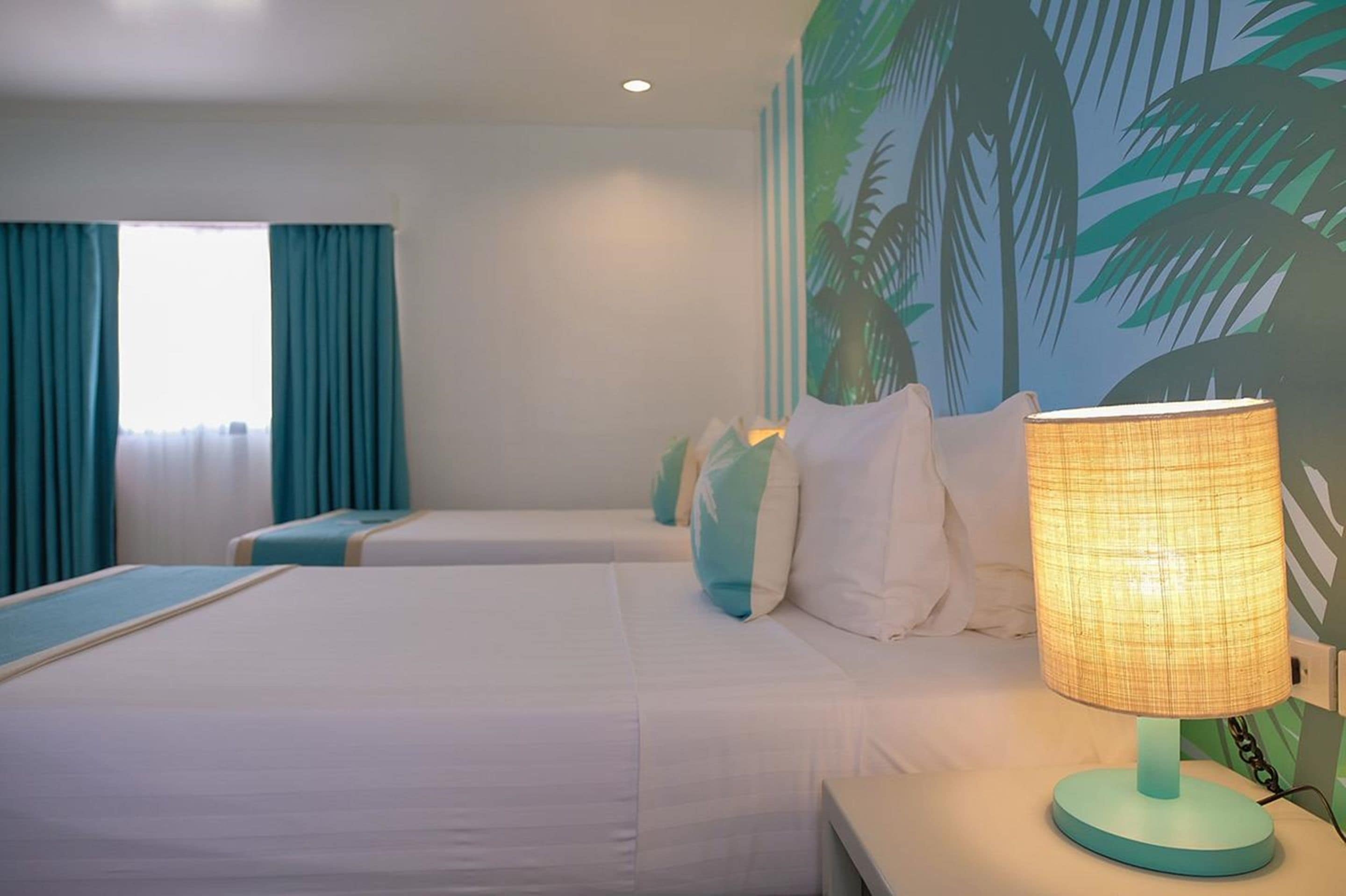 Habitación de hotel con dos camas, cortinas color turquesa, un mural de palmeras y una lámpara encendida.