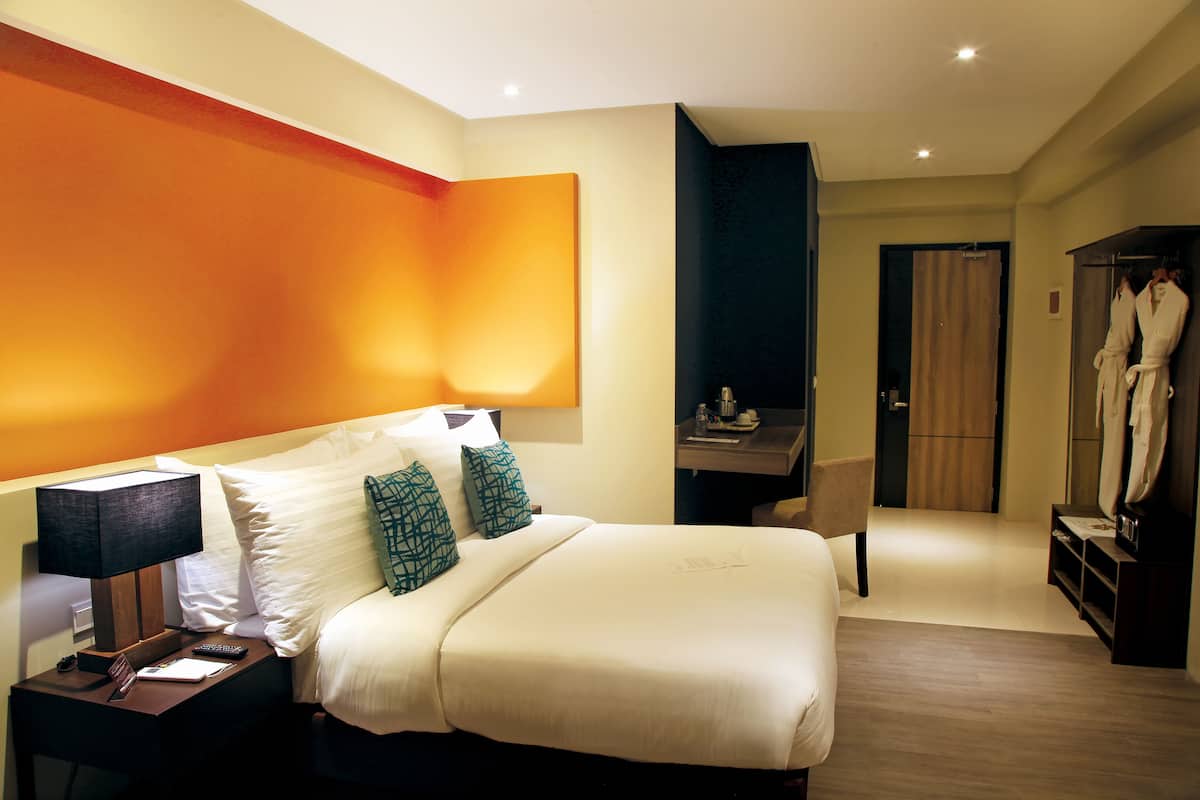 Habitación de hotel con una cama grande, una mesita de noche con lámpara, un escritorio con silla y un armario con batas colgadas.