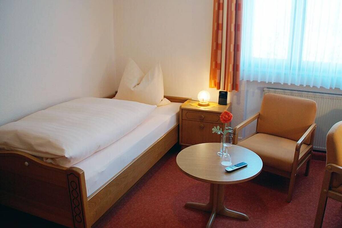A hotel room with a single bed, a bedside table with a lamp, a small round table with a vase and a remote control, and a chair.