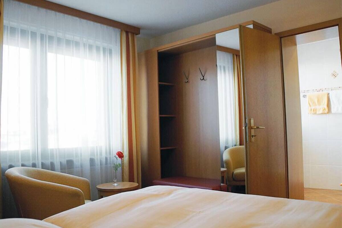 A hotel room with a bed, a chair, a small table with a vase, and a wardrobe.