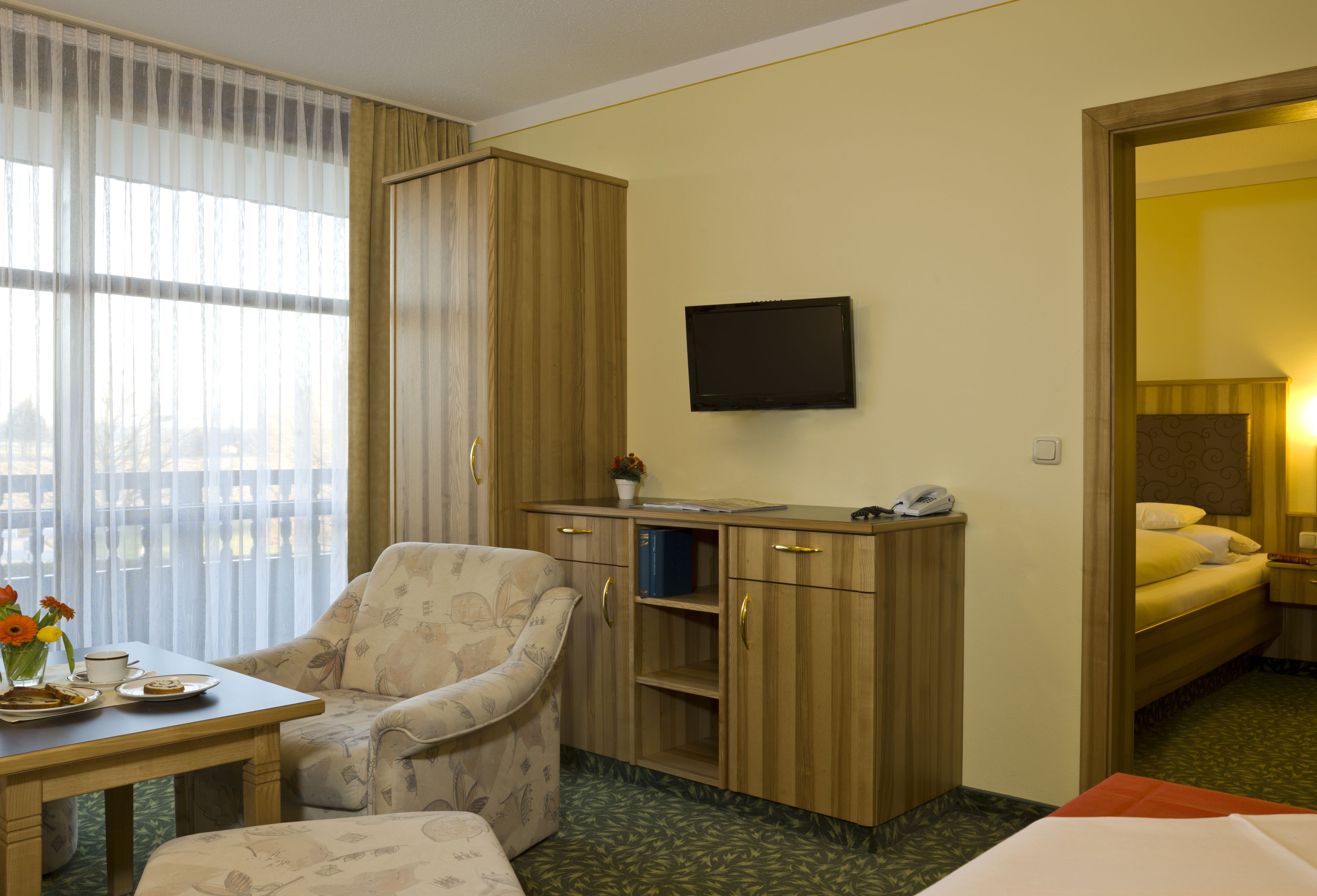 A hotel room with a bed, a desk with a computer, a chair, a TV mounted on the wall, and a window with curtains.