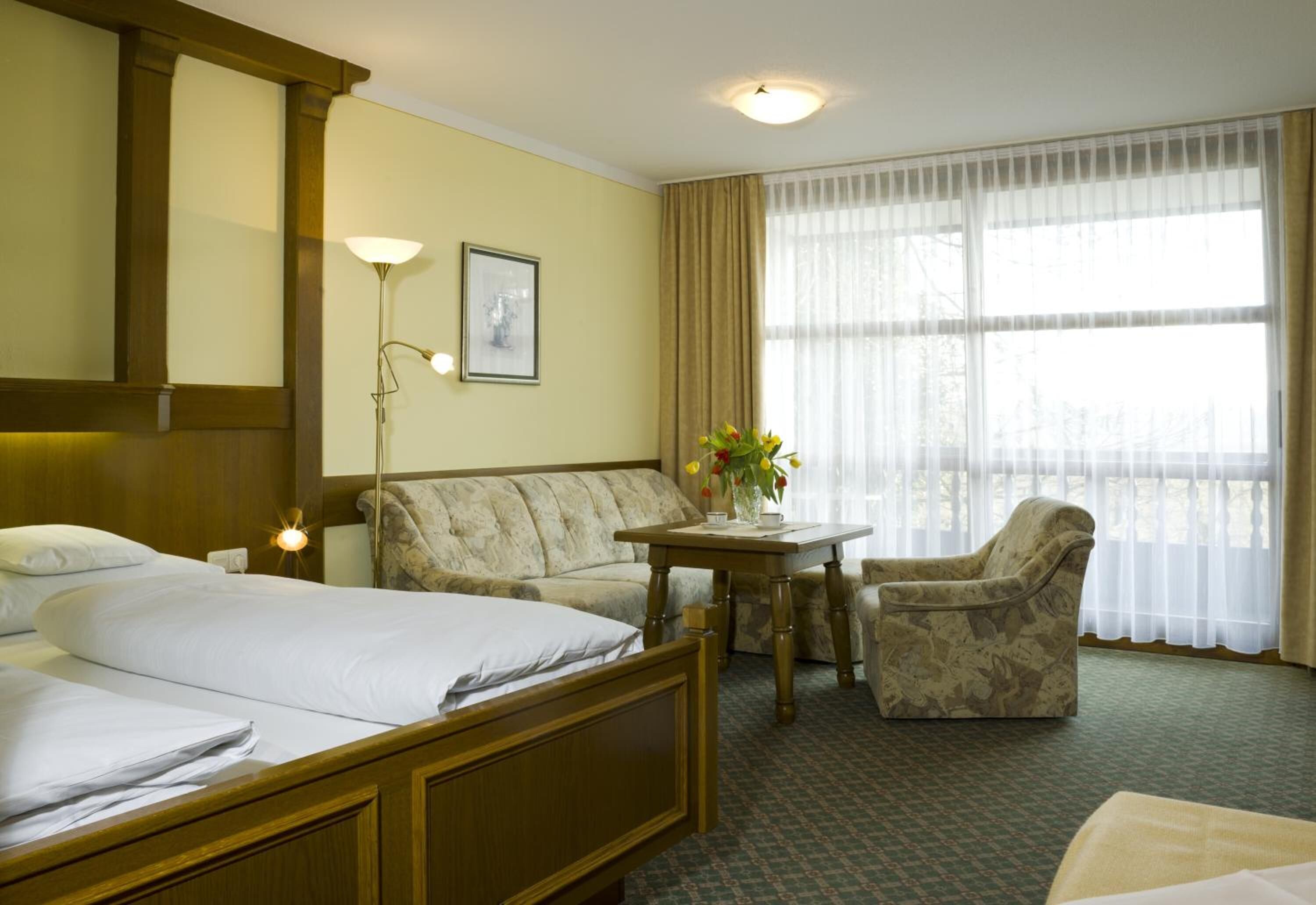 A hotel room with two beds, a sofa, a chair, a table with a vase of flowers, and a large window with curtains.