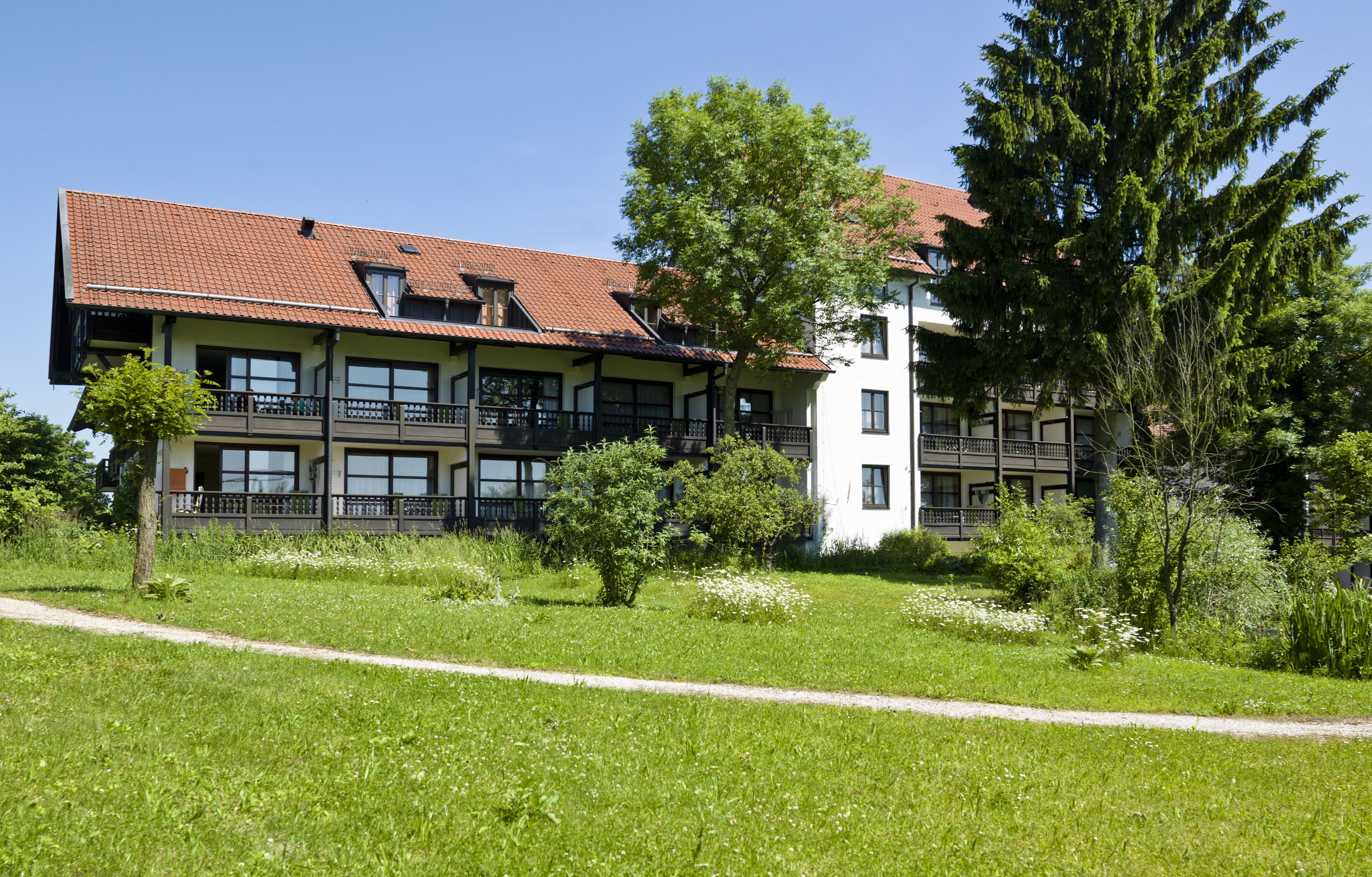 Un immeuble à plusieurs étages, doté d’un toit recouvert de tuiles rouges, de balcons et d’un espace verdoyant avec des arbres.