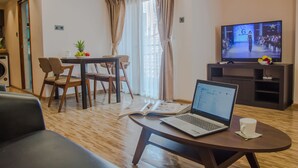 Una sala moderna con una computadora portátil sobre una mesa, un televisor mostrando un desfile de moda y un comedor con sillas y una mesa.