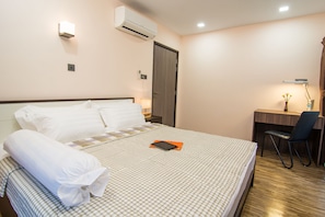 A neatly made bed with pillows, a bedside table with a lamp, a desk with a chair, and an air conditioning unit on the wall.