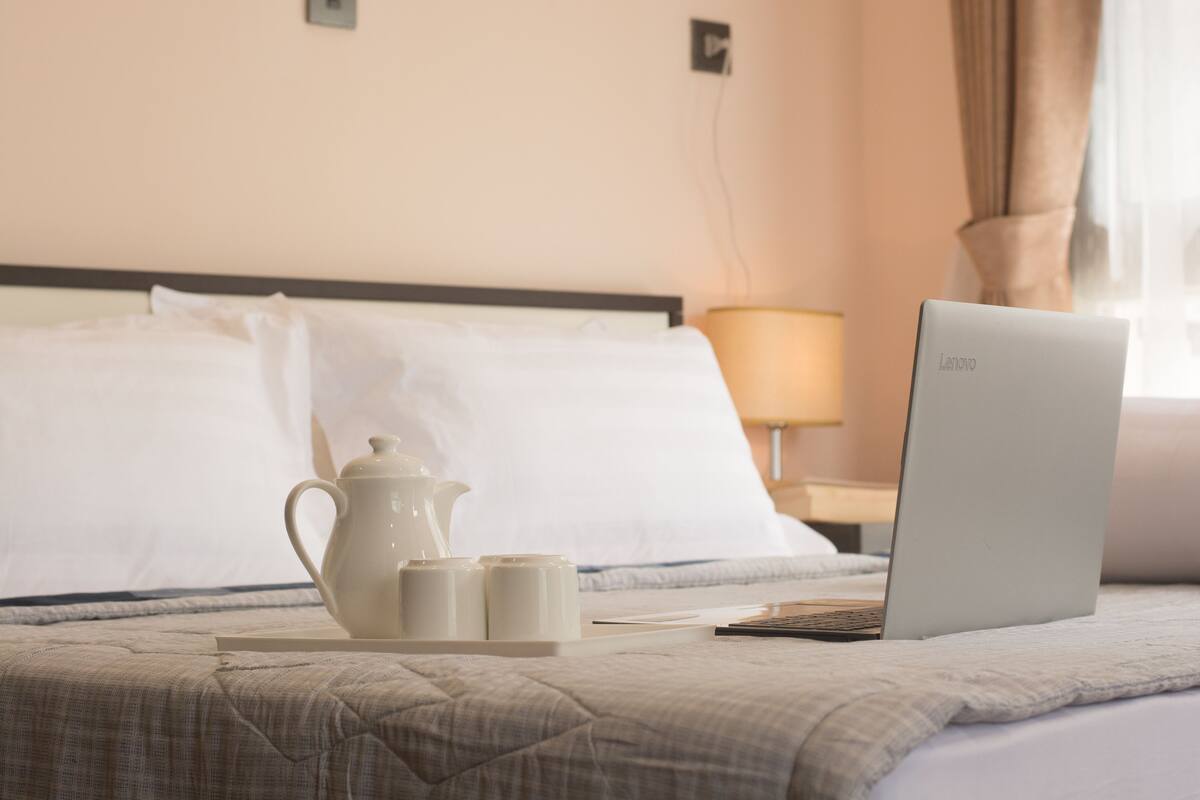 A neatly made bed with a laptop, a teapot, and cups on a tray, suggesting a hotel room setting.
