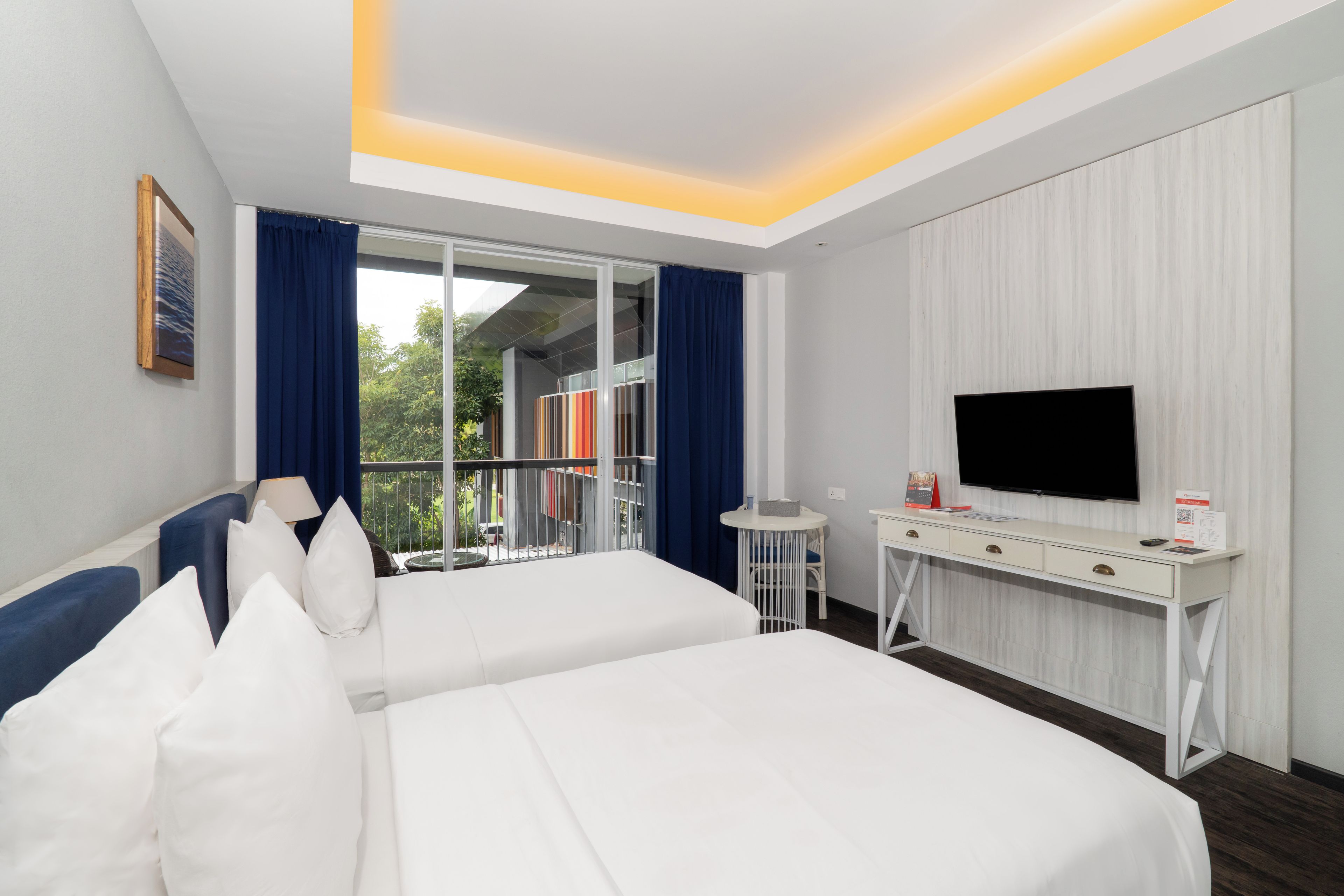 Habitación de hotel con dos camas, un escritorio, un televisor de pantalla plana, una mesita y un balcón con vistas a zonas verdes.