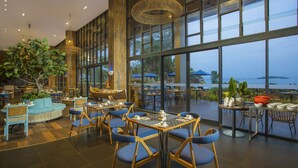 A spacious indoor dining area with large windows, wooden furniture, and a view of the sea.