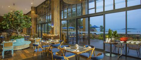 A spacious indoor dining area with large windows, wooden furniture, and a view of the sea.