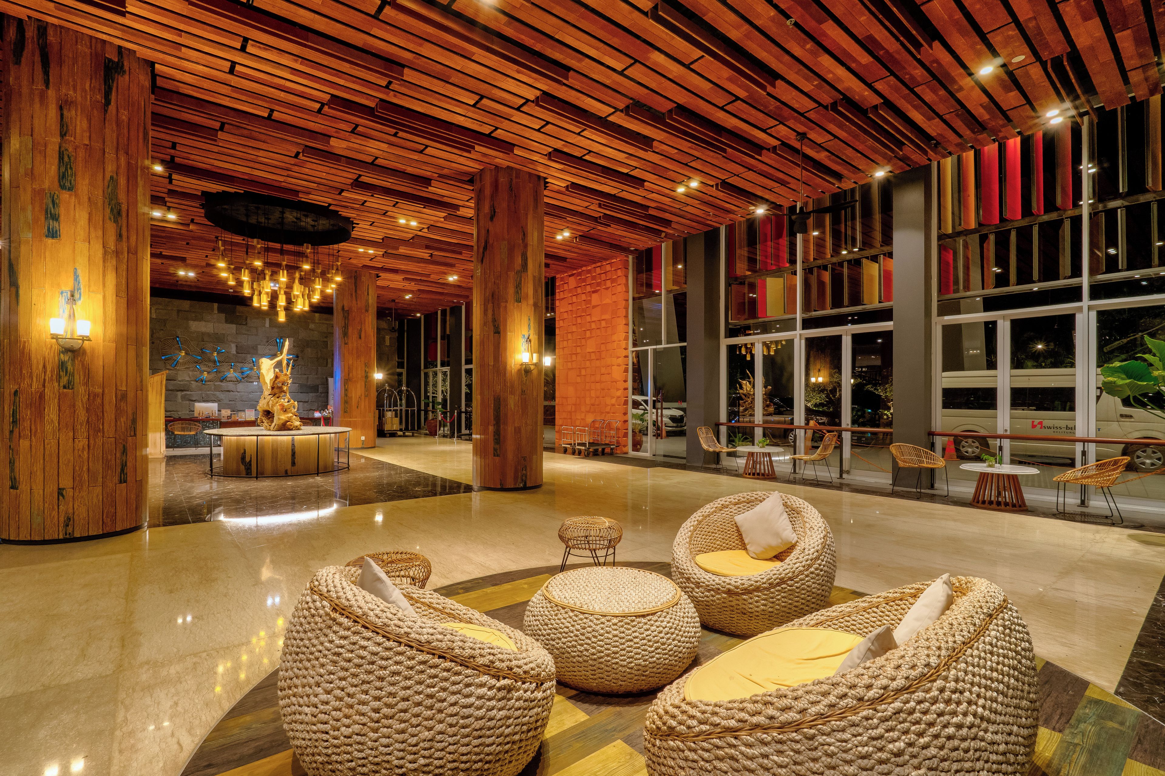 Een ruime lobby met een hoge plafond, houten balken en een centraal zitgedeelte met geweven stoelen.
