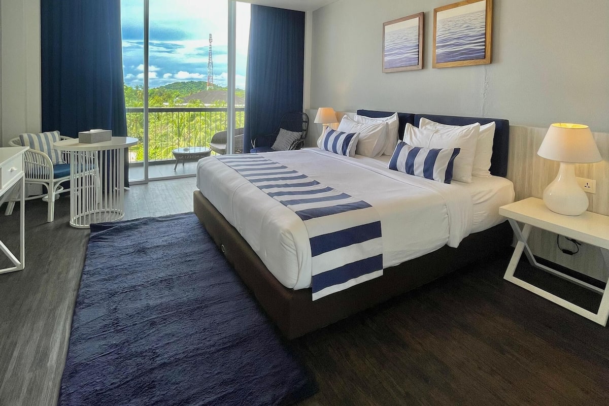 Une chambre d’hôtel moderne, dotée d’un grand lit, d’un bureau et d’une chaise. Les fenêtres offrent une vue sur l’extérieur.