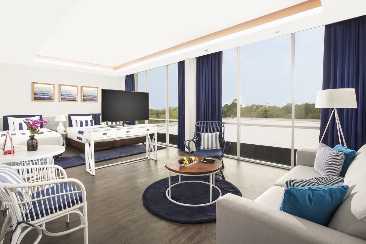 Une chambre d’hôtel moderne dotée d’une grande fenêtre, d’un téléviseur à écran plat, d’un coin repas avec une table ronde et d’un canapé agrémenté de coussins bleus.