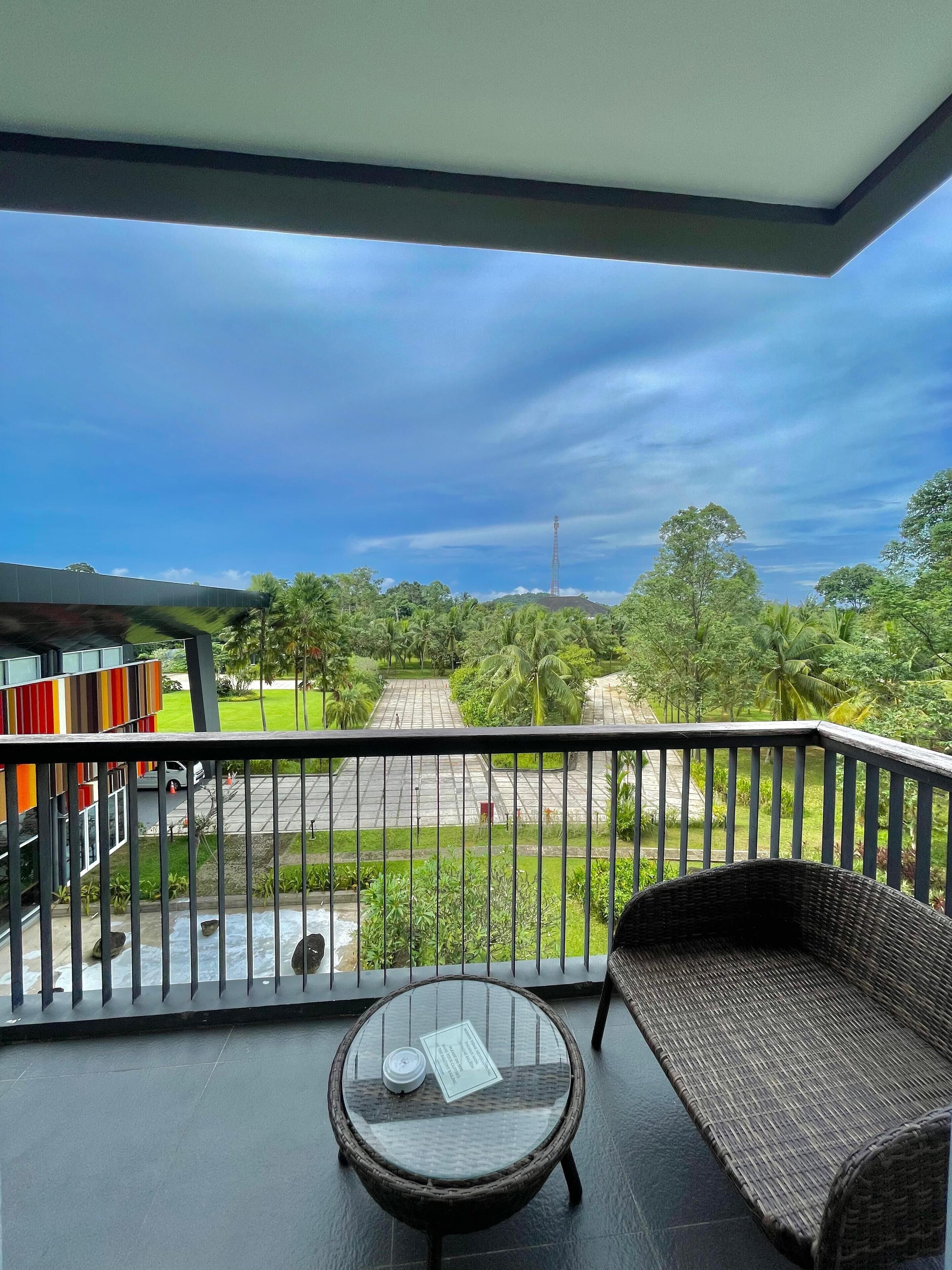 A balcony with a wicker sofa and a glass table, overlooking a paved area with greenery and a distant tower.