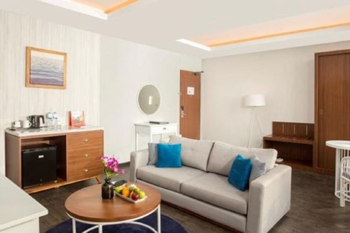 Une chambre d’hôtel moderne avec un canapé, une petite table ronde sur laquelle sont disposés des fruits, une machine à café et une vue sur la mer par la fenêtre.