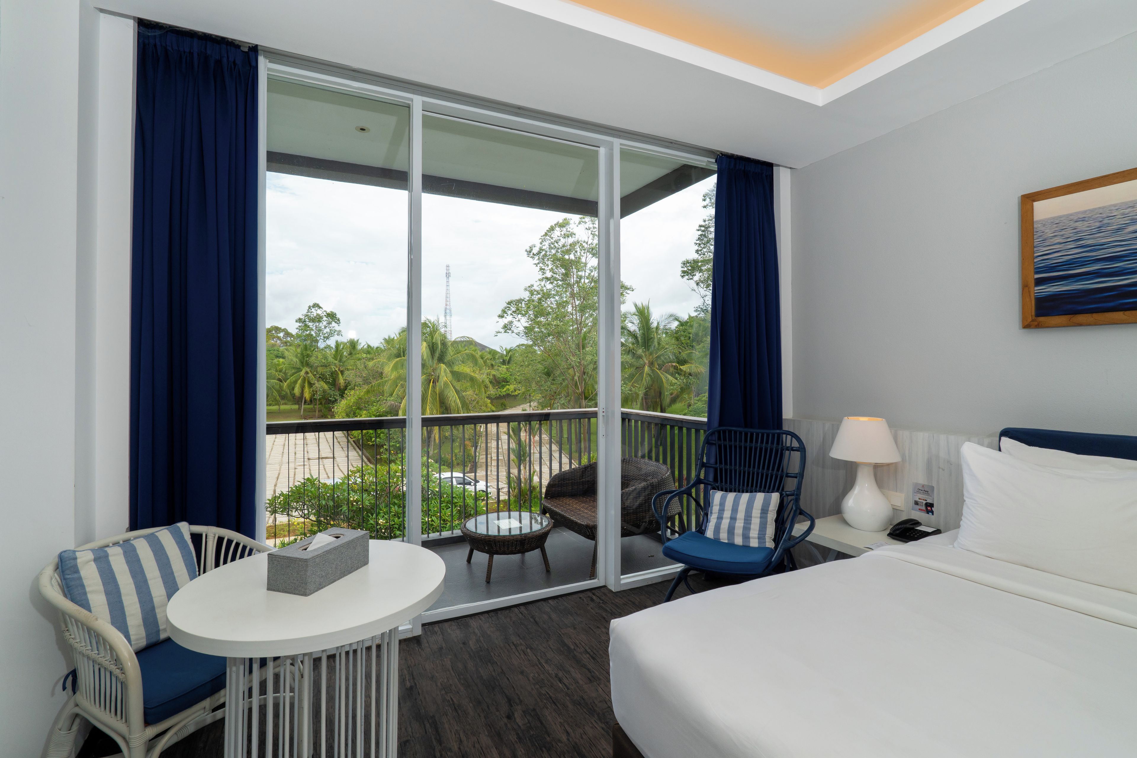 Ein Hotelzimmer mit einem Bett, einem kleinen Tisch, zwei Stühlen, einer Lampe und Blick auf Grün durch große Fenster.