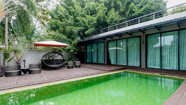A swimming pool with a wooden deck, surrounded by a glass wall and a covered seating area with chairs and an umbrella. - The Kris BangTao by Lofty (Choeng Thale)