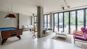 A spacious living area with a pool table, a sofa, a coffee table, and a view of the outdoors through large windows. - The Kris BangTao by Lofty (Choeng Thale)