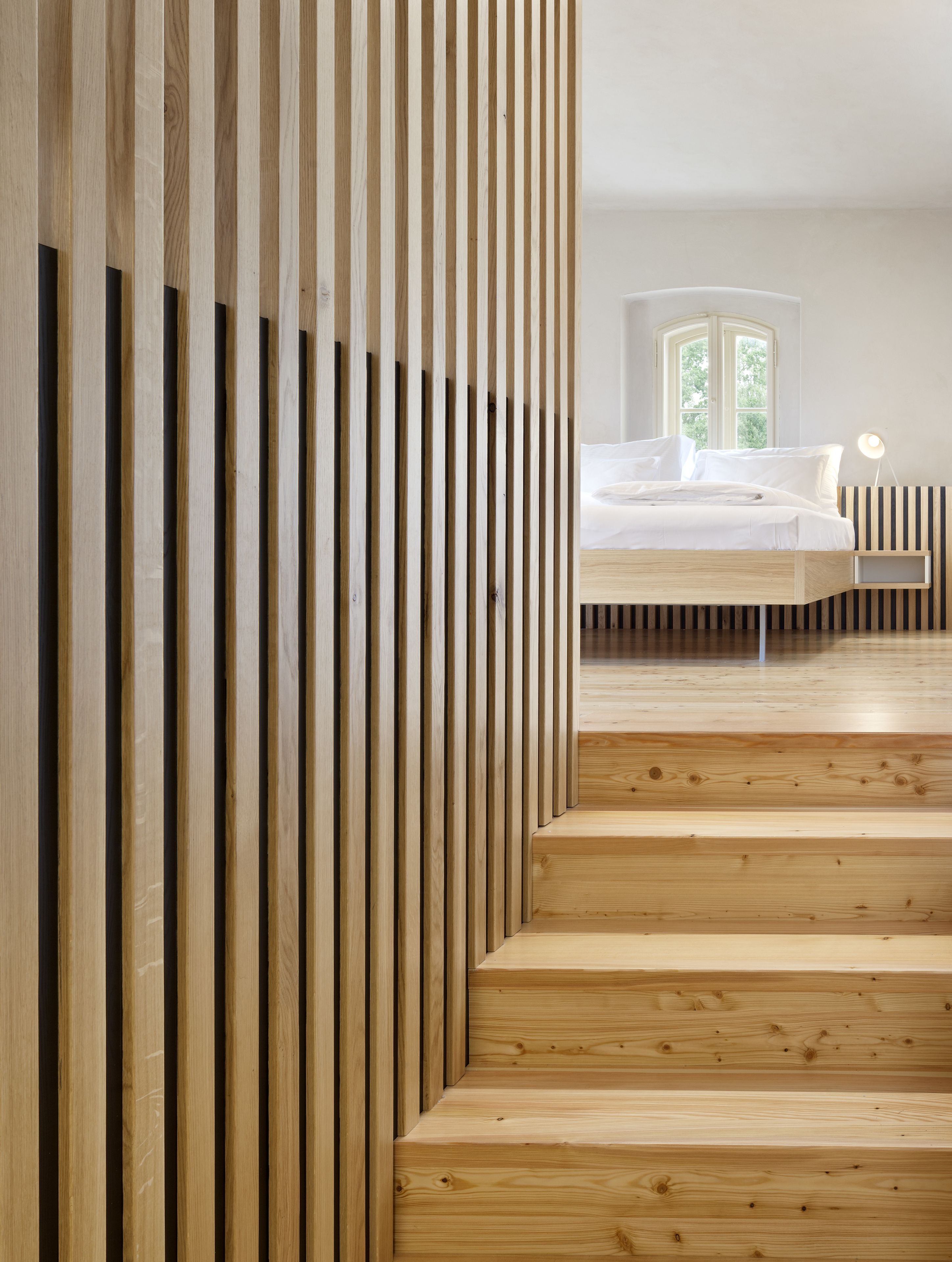 Camera da letto moderna con una scala in legno che conduce a un letto, una finestra con vista su piante e una lampada da comodino.