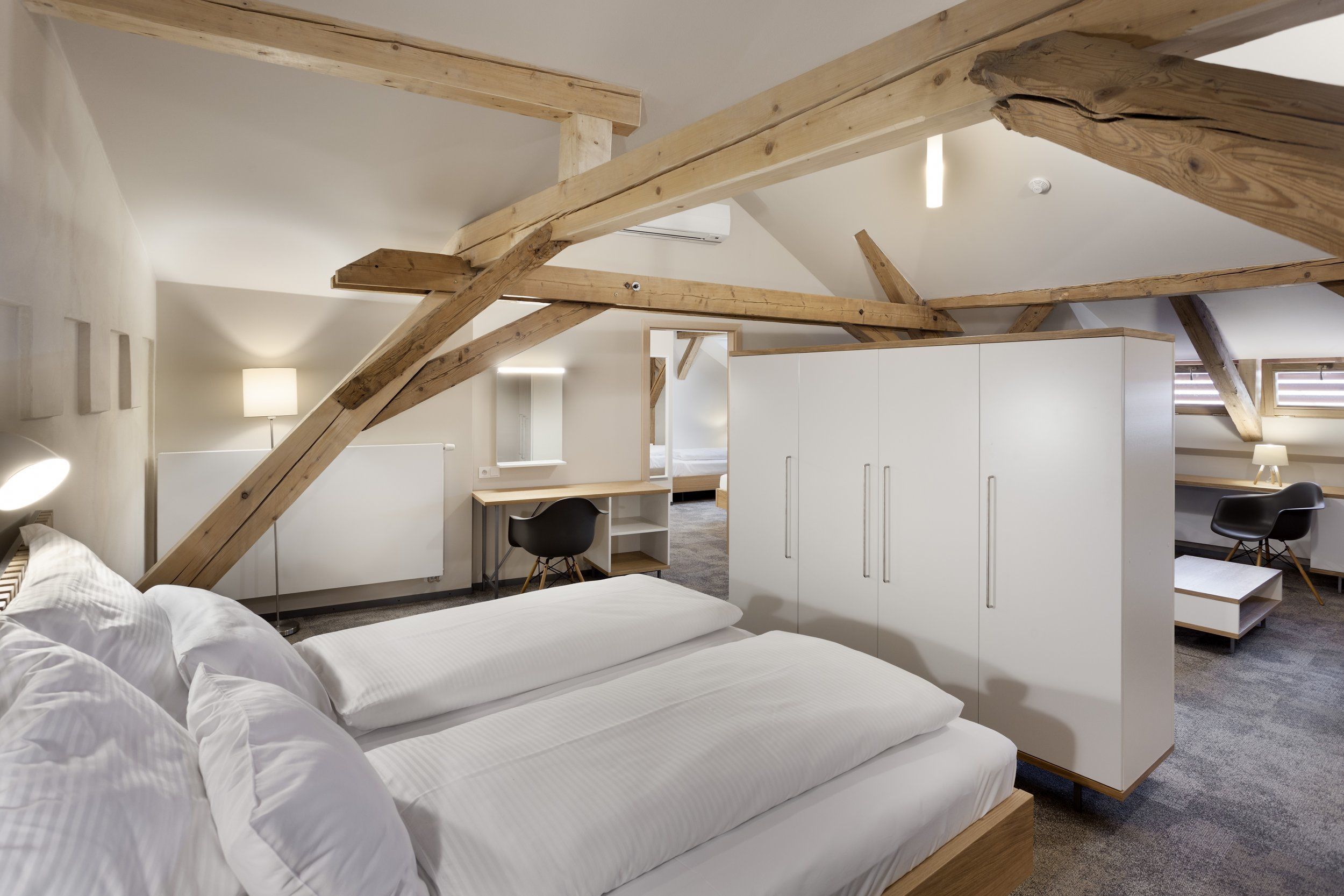 A modern loft bedroom with exposed wooden beams, a desk, and a wardrobe.