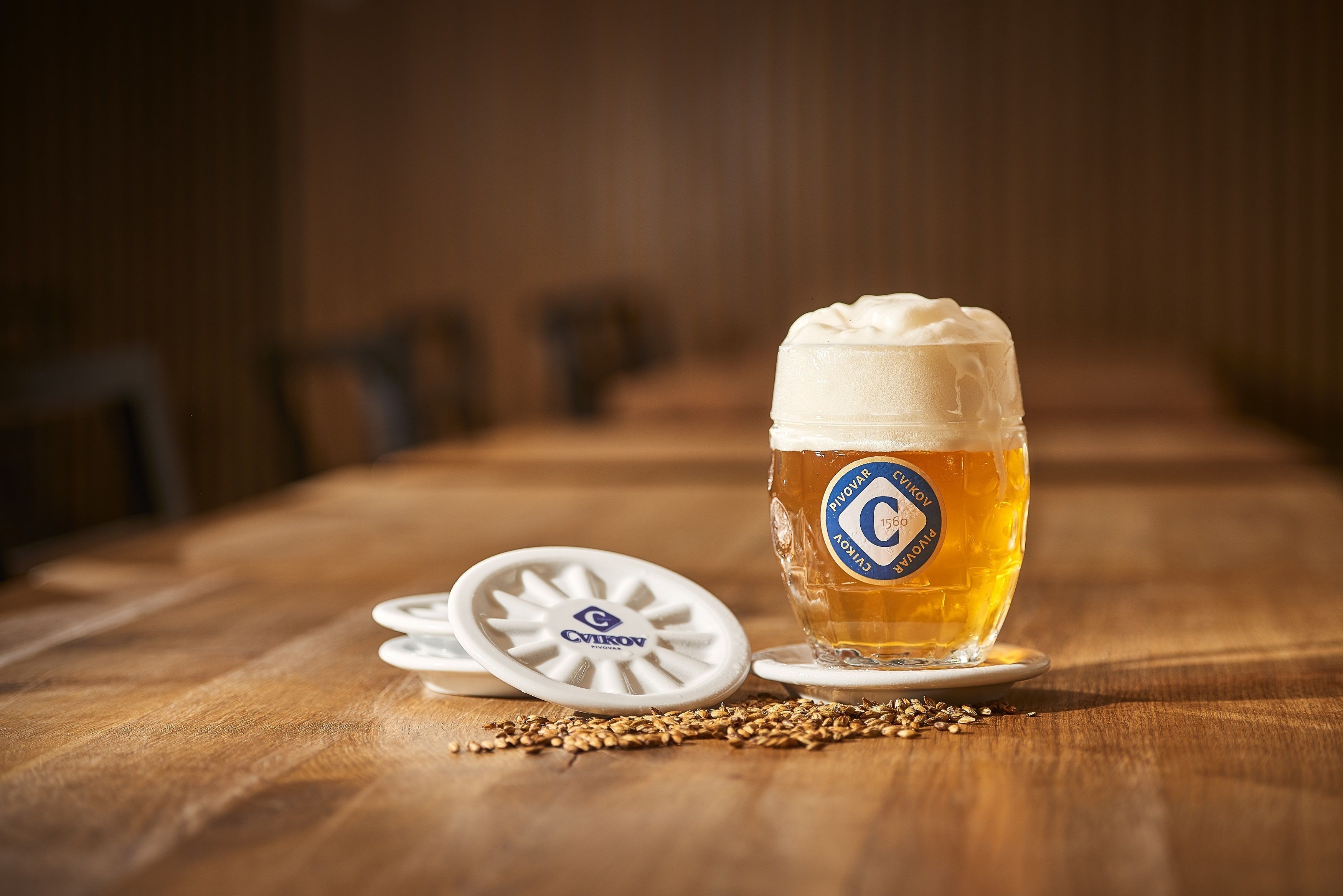 Una cerveza en un vaso con una etiqueta sobre un posavasos, sobre una mesa de madera con granos esparcidos.