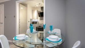 A modern dining area with a glass table and white chairs, a kitchen in the background, and a door leading to another room. - Forbes Retreat - Donnini Apartments (Ayr)