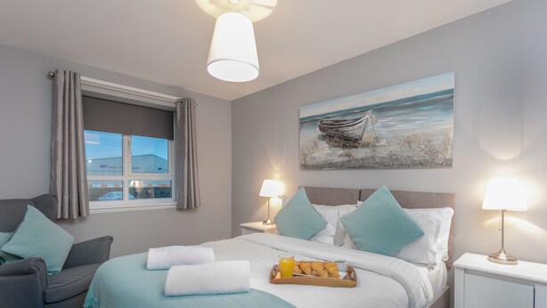 A hotel room with a bed, a chair, a nightstand, and a painting of a boat on the wall. - Forbes Retreat - Donnini Apartments (Ayr)