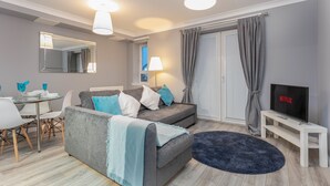 A modern living room with a grey sofa, a TV on a white stand, a dining table with chairs, and a large mirror on the wall. - Forbes Retreat - Donnini Apartments (Ayr)