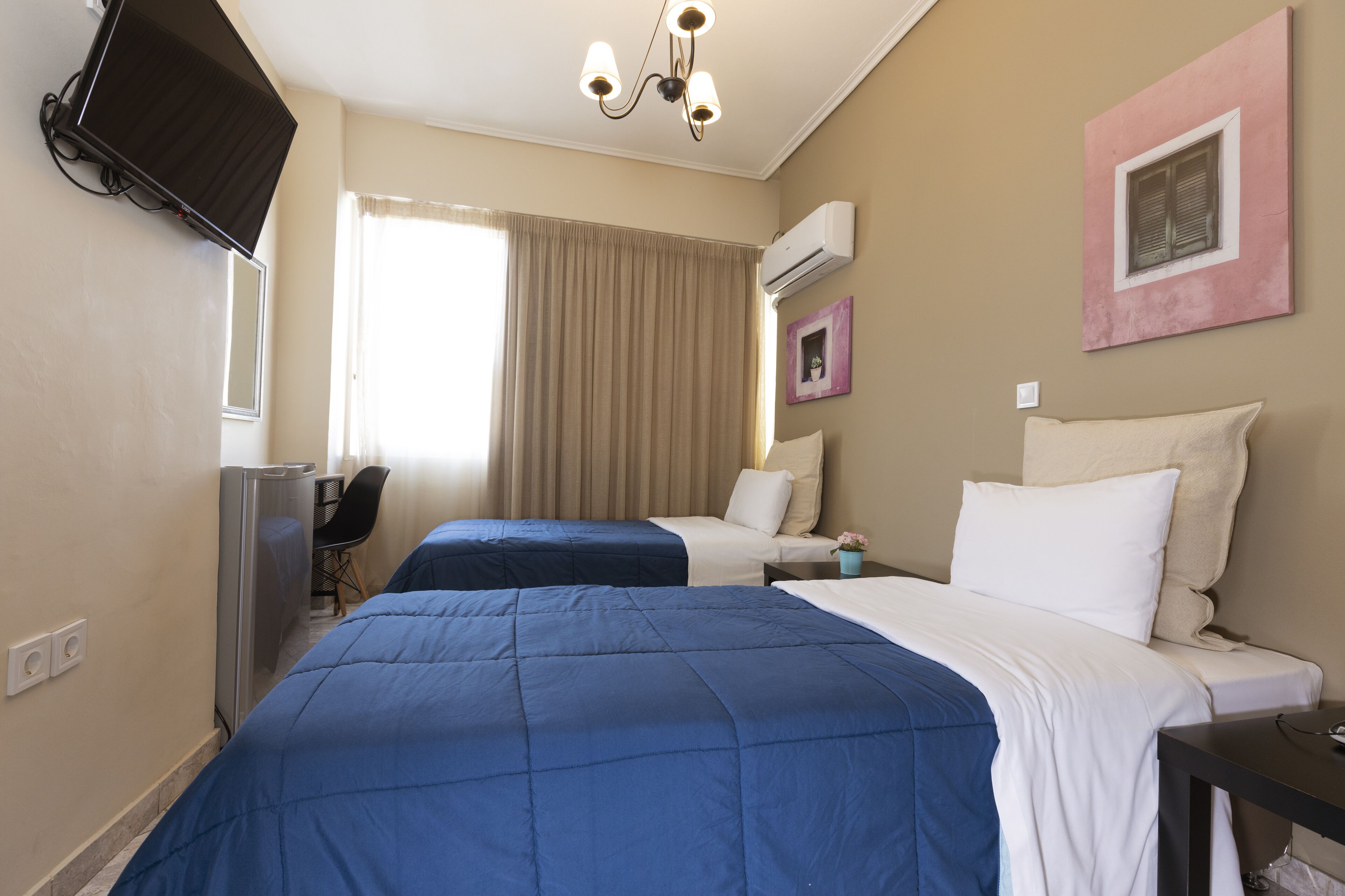 A hotel room with two beds, a desk, a chair, a TV, and an air conditioning unit.