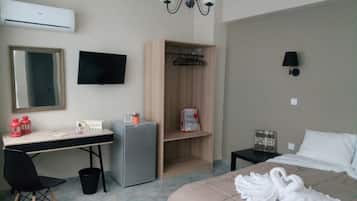 A hotel room with a bed, a desk with a computer, a small refrigerator, a mirror, a TV, a wardrobe, and a bedside table with a lamp.