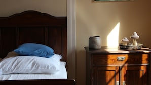 Een keurig opgemaakt bed met witte lakens en een blauw kussen, een houten dressoir met een metalen slot, en een vaas met decoratieve objecten erop.