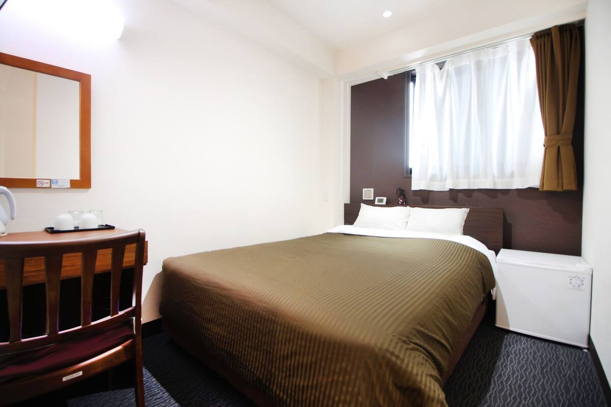 A hotel room with a bed, a desk with a chair, a small refrigerator, and a window with curtains.