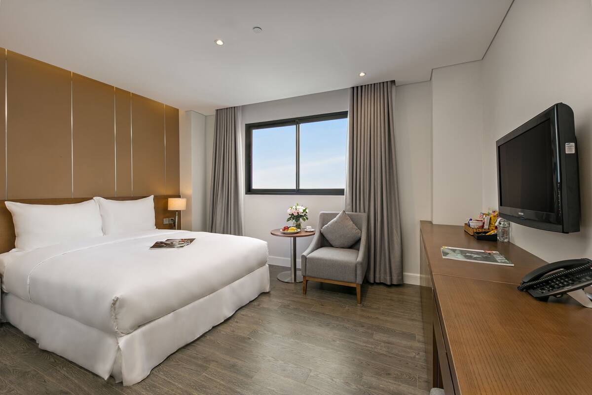 A hotel room with a large bed, a small table with a vase of flowers, a chair, a desk with a phone, and a TV.