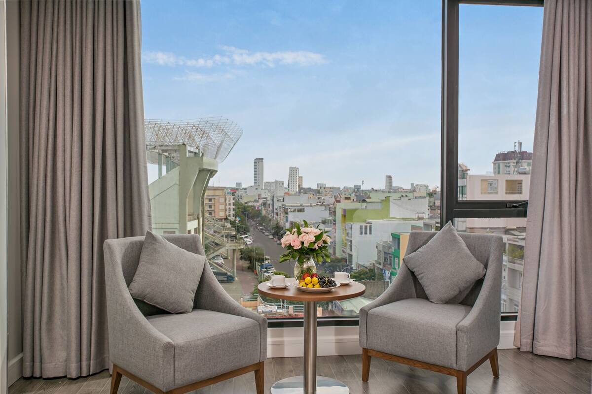 A modern hotel room with two grey armchairs, a small round table with a vase of flowers, and a city view through large windows.