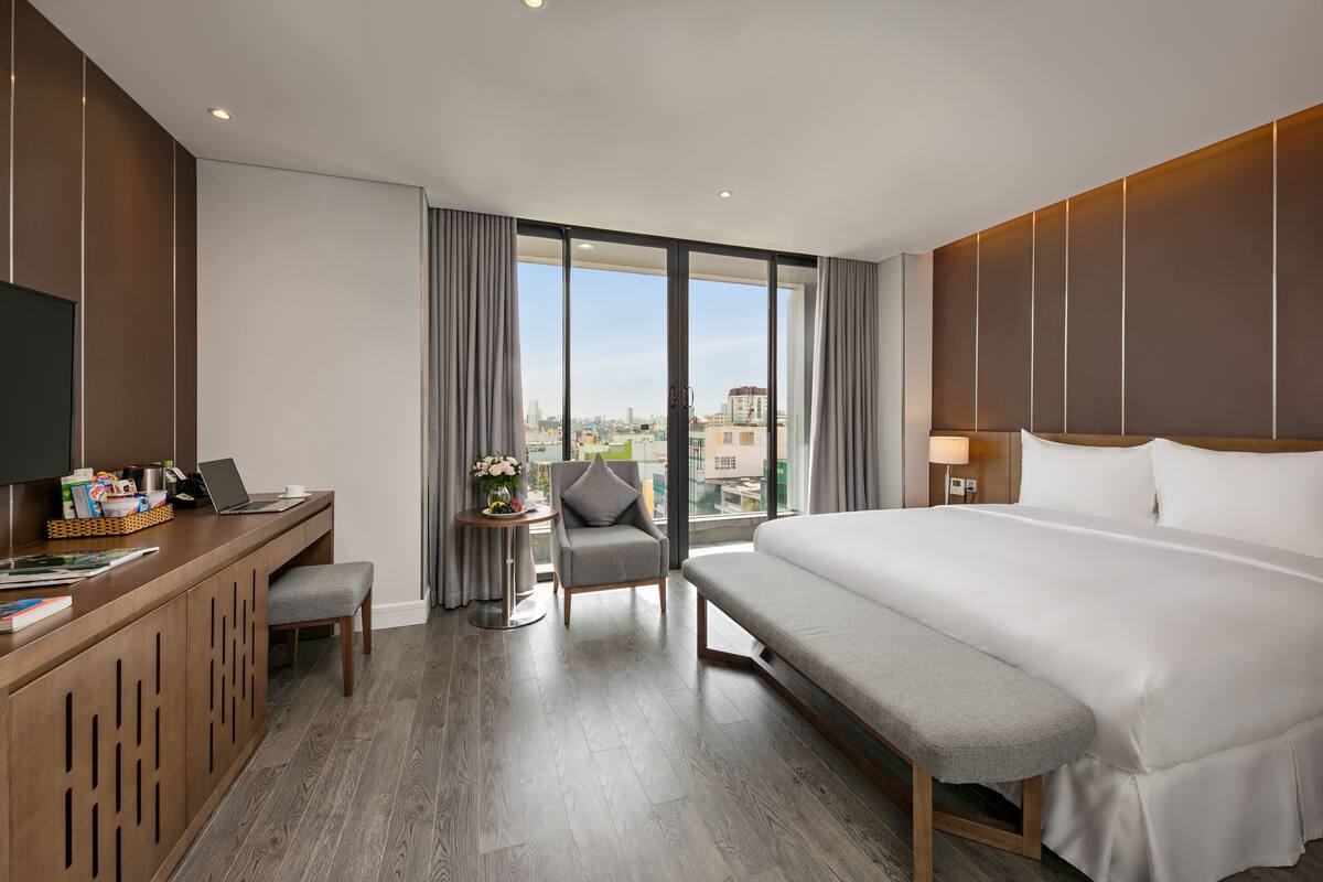A modern hotel room with a large bed, a desk with a laptop, a chair, and a view of the city through large windows.