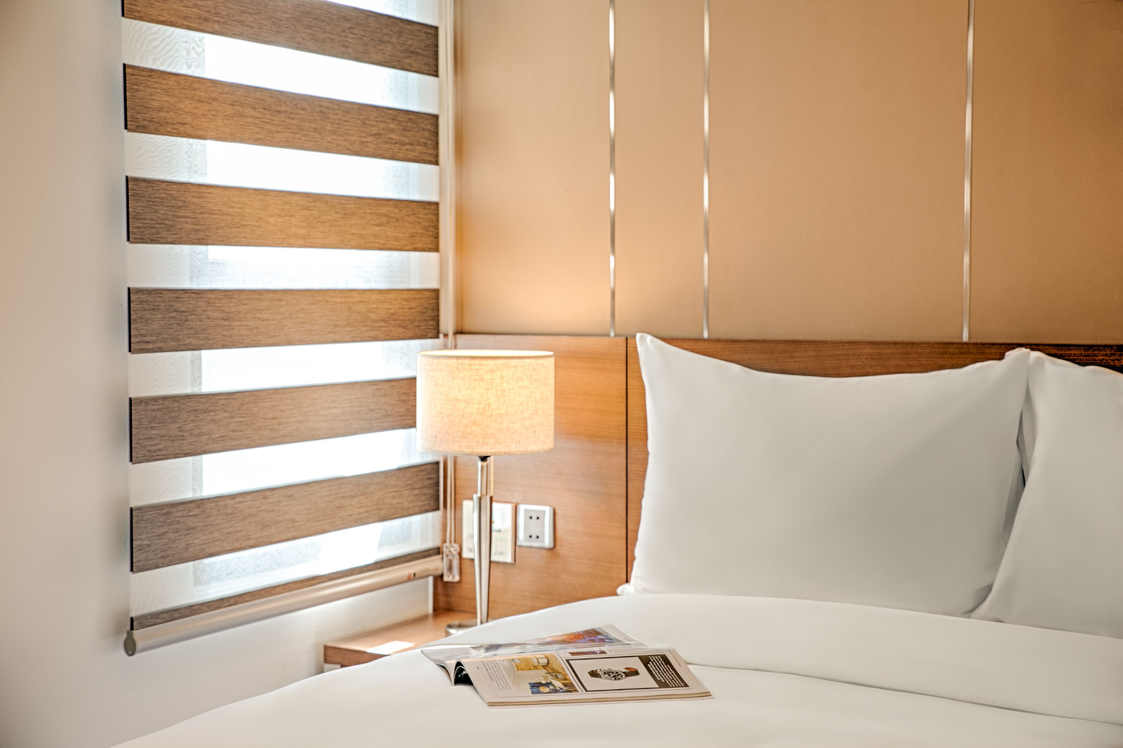Habitación de hotel con cabecera de madera, lámpara de noche, cama blanca y una revista sobre la cama.