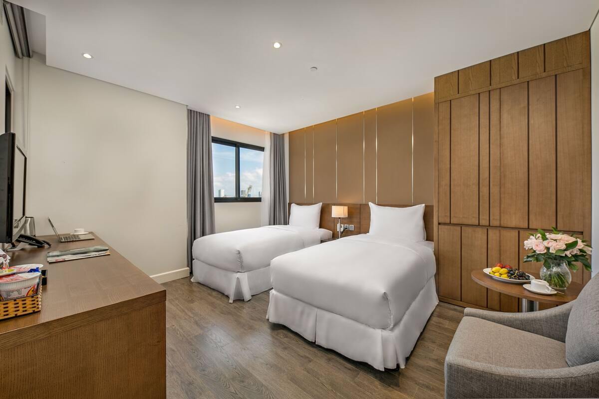 A hotel room with two beds, a desk with a computer and phone, a chair, a small table with a vase of flowers, and a window with curtains.