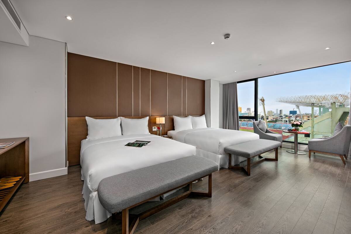 A modern hotel room with two beds, a bench, a desk, and a view of the city.