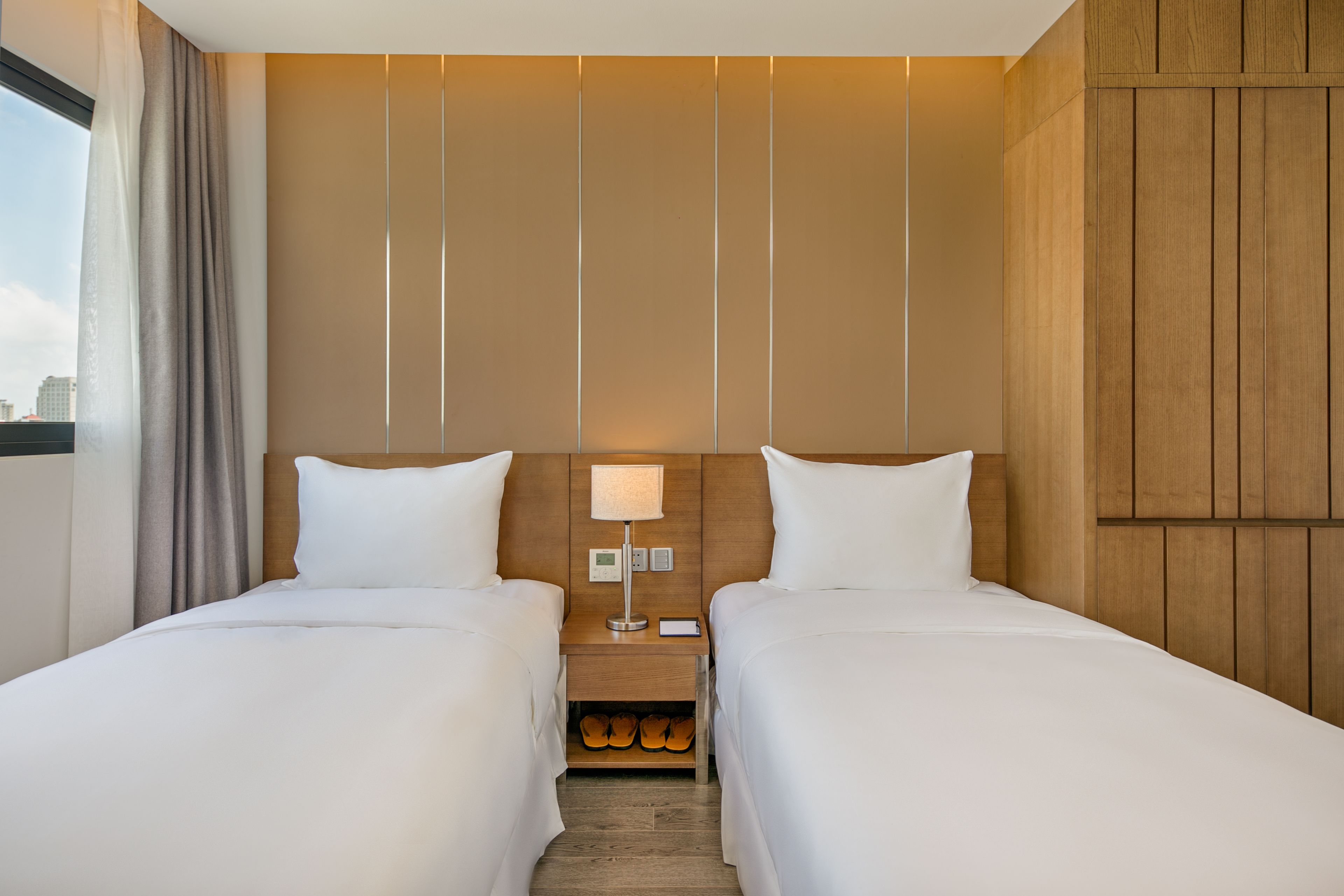 Habitación de hotel con dos camas, una mesita de noche, una lámpara y espacio para guardar zapatos.
