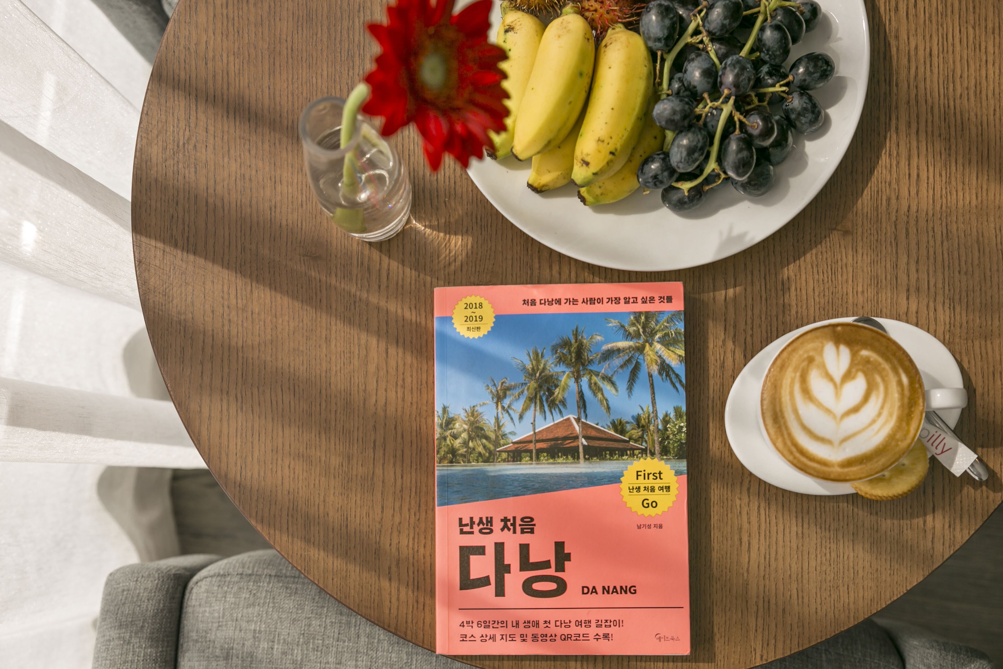 A round wooden table with a plate of bananas and grapes, a book titled 'DA NANG', and a cup of coffee with latte art.