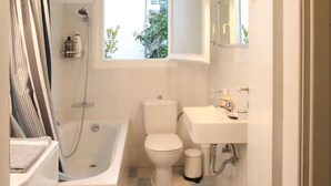 A compact bathroom with a bathtub, toilet, and sink, featuring a checkered floor and a window with a view of greenery outside. - Stylish flat by the beach of Edem (Palaio Faliro)