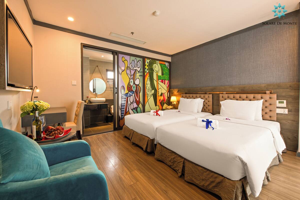A hotel room with two beds, a sofa, a dining table with a vase of flowers, and a colorful mural on the wall.