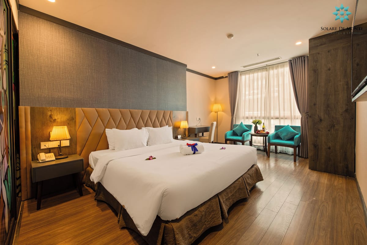 A hotel room with a large bed, a bedside table with a lamp, a small table with a plant, and a seating area with chairs.