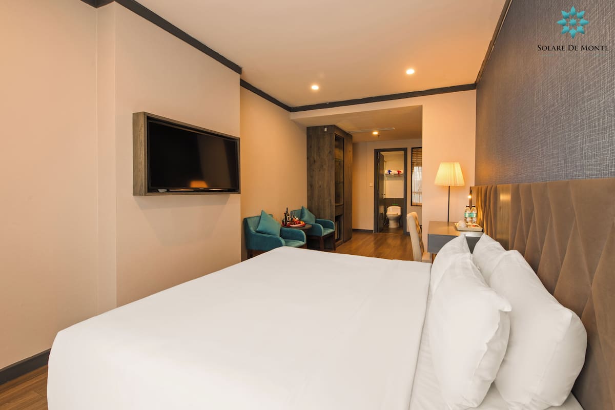 A hotel room with a large bed, a flat-screen TV mounted on the wall, a seating area with two chairs, and a lamp on a side table.