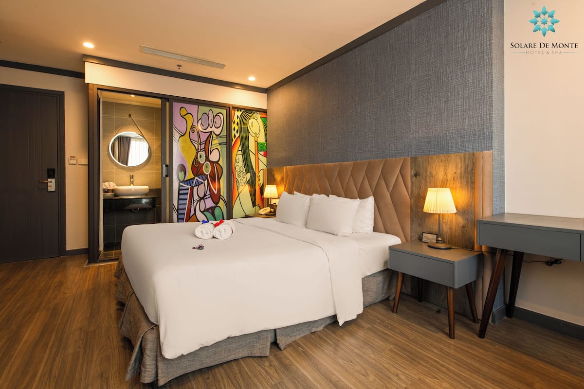 A modern hotel room with a large bed, a bedside table with a lamp, a small desk, a round mirror, and a colorful abstract painting on the wall.