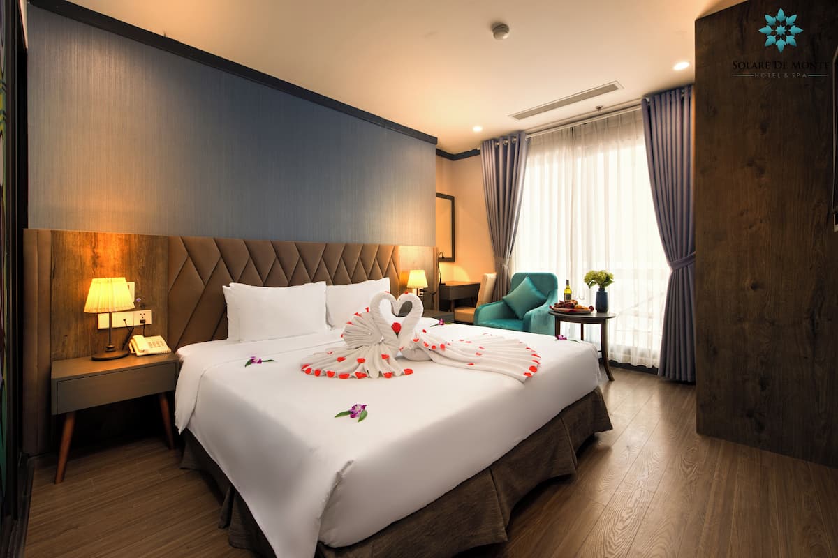 A hotel room with a large bed, a wooden headboard, a nightstand with a lamp, a small table with a plant, and a teal chair.