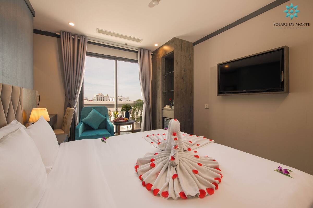 A hotel room with a large bed, a TV on the wall, a view of the city through the window, and a chair by the window.